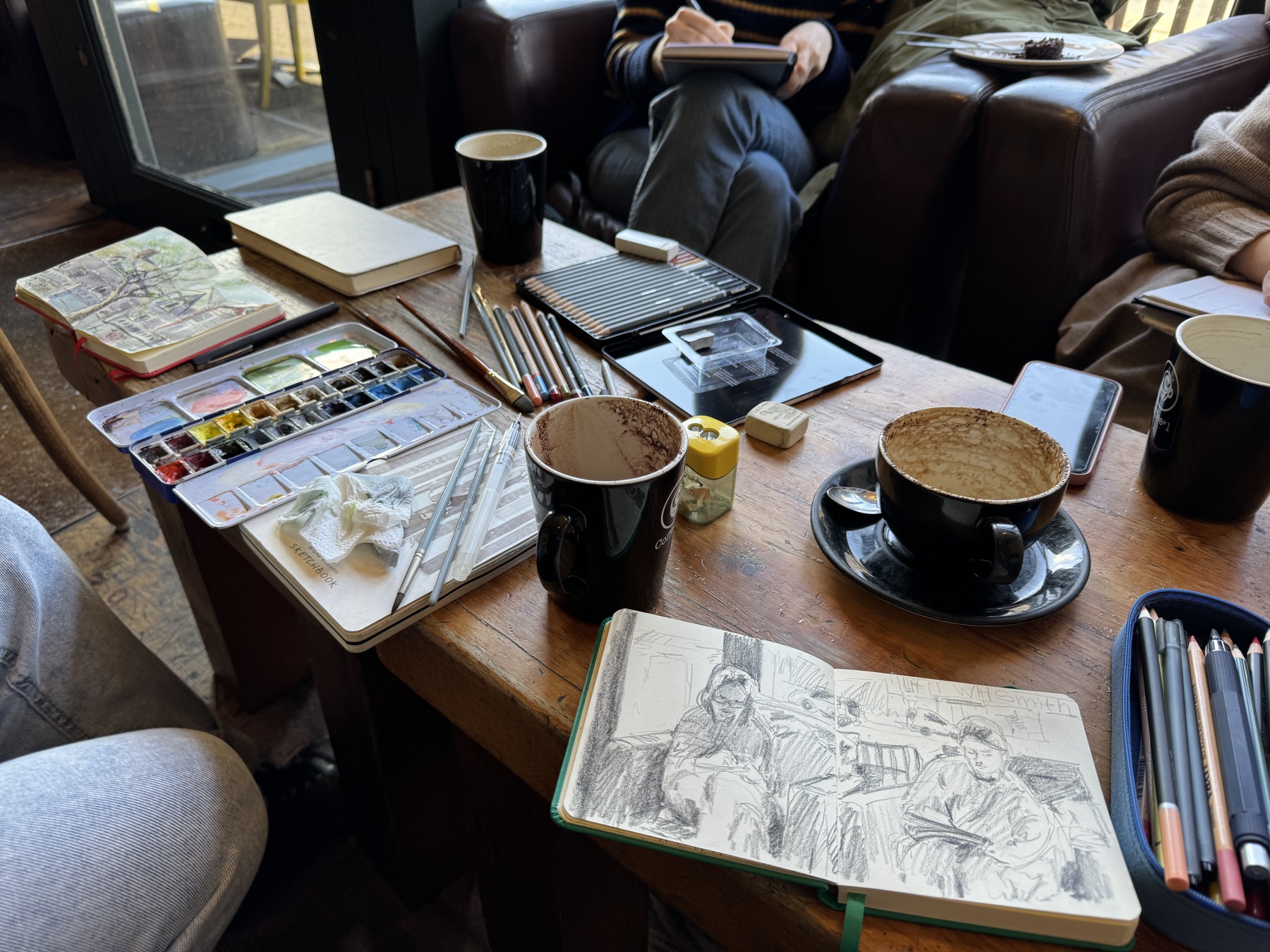 Cafe table with sketchbooks, pencils, watercolours, and coffee cups