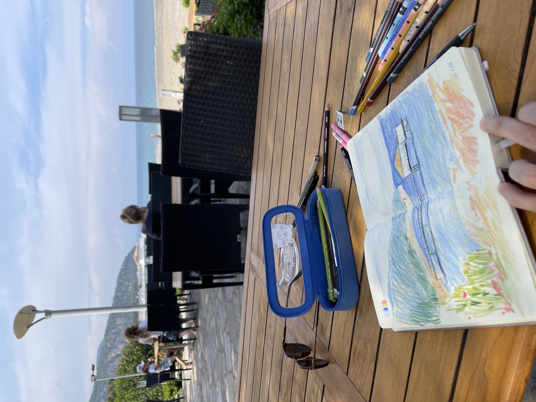 Sketchbook and coloured pencils on a table with a sea view beyond
